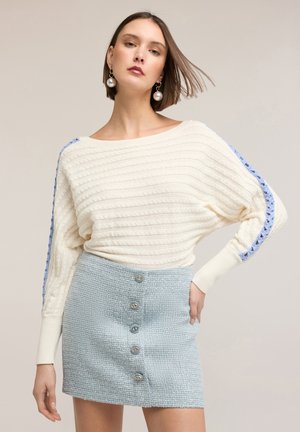 Junge Frau mit kurzem braunem Haar trägt einen weißen Strickpullover mit blauem Streifen und einen hellblauen, strukturierten Rock mit Knöpfen.