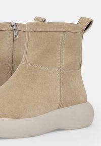 Beige mokasini nilkkurit, joissa on pyöreä beige pohja, sivuvetoketju ja takana kätevä lenkki pukemista helpottamaan.