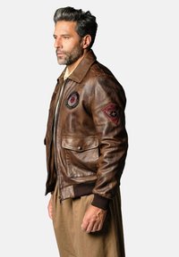 Veste aviateur en cuir marron avec fermeture éclair sur le devant, poignets côtelés, deux écussons sur la poitrine, grandes poches et coupe structurée. Détail de surface texturée.