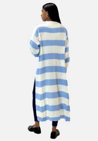 Lång cardigan i tjock stickning med horisontella blå och vita ränder. Har ribbad textur och midi-längd. Bärs över mörka jeans.