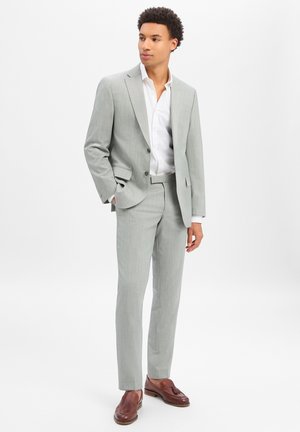 Mann trägt hellgrauen Anzug mit weißem Hemd und braunen Loafers, steht mit einer Hand in der Tasche und schaut seitlich vor weißem Hintergrund.