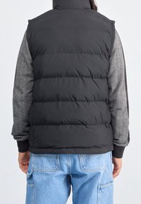 Schwarze gepolsterte Weste mit hohem Kragen, die eine glatte Textur und eine taillierte Passform aufweist, getragen über einem grau gemusterten Langarmshirt und blauen Jeans.