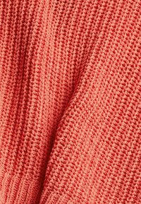 Sweter dziany w kolorze koralowym, przedstawiający teksturowany wzór z widocznymi ściegami oraz miękką, oddychającą tkaniną.