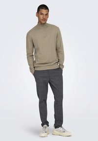 Pull beige à manches longues avec un col montant, assorti à un pantalon gris à motif à carreaux. Le modèle se tient devant un fond neutre, portant des baskets gris clair.