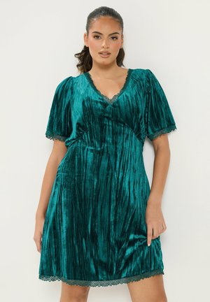 Femme portant une robe en velours sarcelle avec des manches courtes bouffantes et une bordure en dentelle sur le décolleté, les manches et l'ourlet, debout devant un fond uni.