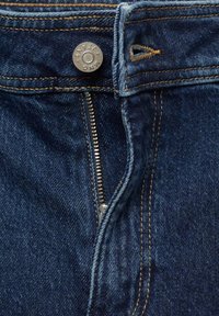 Gros plan sur un jean en denim bleu foncé avec une fermeture éclair métallique partiellement ouverte et un bouton métallique étiqueté "MNG JEANS."