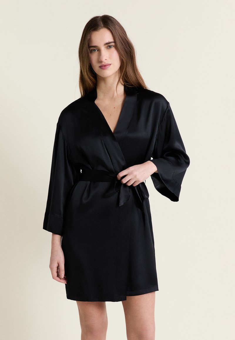 Jeune femme portant une robe portefeuille en satin noir, en train de nouer la ceinture, debout devant un fond beige uni.