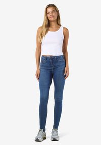Bílý žebrovaný top bez rukávů v kombinaci s modrými úzkými džíny. Modelka má na sobě šedé tenisky. Jednoduchý design, přiléhavý střih, hladká textura.