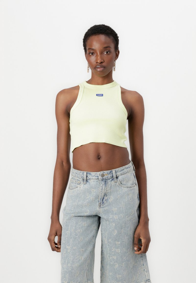 Ljusgrön ribbad crop top med hög krage, som visar en logotyp på bröstet. Bärs tillsammans med ljusblå vida denimjeans.