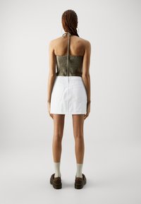 Haut à bretelles smocké couleur olive avec un lien à l'arrière, associé à une mini-jupe en denim blanche avec poches et coupe droite. Chaussures compensées marron visibles.