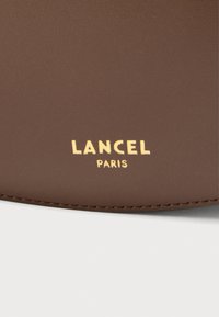 Produs din piele maro cu text auriu embossat „LANCEL PARIS” în partea de jos; textură netedă cu cusături vizibile de-a lungul marginilor.
