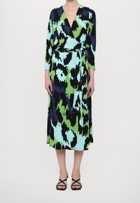 Zavinovací šaty v modrém, zeleném a černém abstraktním vzoru; dlouhé rukávy a zavazování v pase; střední délka; v kombinaci s černými sandály na podpatku.