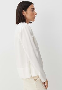 Camicia bianca con maniche lunghe, tessuto morbido, collo rotondo e orlo leggermente asimmetrico. Texture liscia con polsini abbottonati.