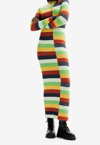 Personne debout les bras croisés portant une robe maxi côtelée rayée colorée et des bottes de combat noires à lacets sur fond blanc.