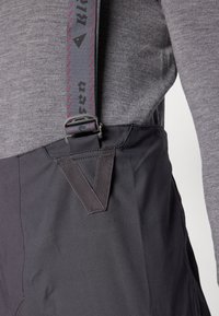 Graues Langarmshirt mit schwarzen verstellbaren Hosenträgern. Die Hosenträger haben rote Punkte und eine Metallklammer, die am grauen Stoff des Shirts befestigt ist.