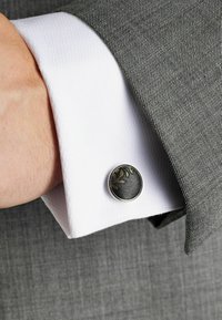 Auksinė lapų rašto apdaila suapvalinto juodo raštuoto paviršiaus manžetų sagė, pritvirtinta prie baltos tekstūruotos dress shirt manžetės. Pilkas kostiumo audinys fone.