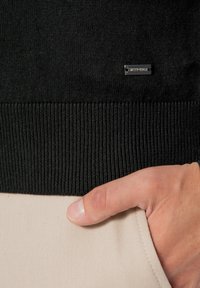 Schwarzer Strickpullover mit geripptem Saum, ausgestattet mit einem kleinen silbernen Logo-Tag. Hand ruht auf einer hellen Tasche. Weiche Textur.