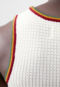Hvit strikket tank topp med et teksturert mønster, som har flerfargede stripete detaljer i rødt, grønt og gult langs halsen.