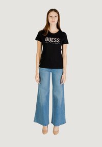 T-shirt nera di cotone con "GUESS LOS ANGELES" stampato in lettere bianche in grassetto, abbinata a jeans a zampa d'elefante blu chiaro e tacchi beige.