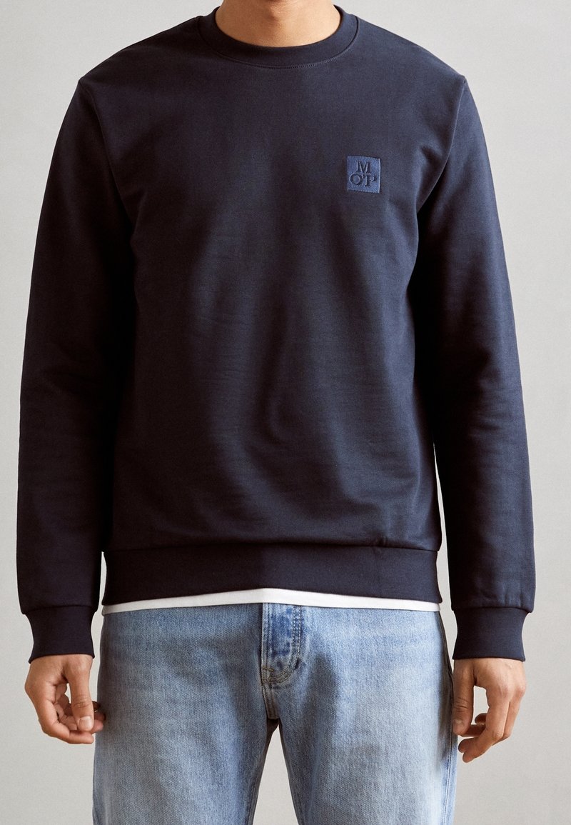 Homme portant un sweat-shirt bleu marine foncé avec le logo MOP sur la poitrine, un jean bleu clair et une chemise blanche dépassant sous l'ourlet du sweat-shirt.