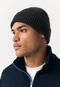 Dark ribbed knit beanie, round shape, worn above the forehead; accompanied by a dark zip-up jacket and white t-shirt underneath.