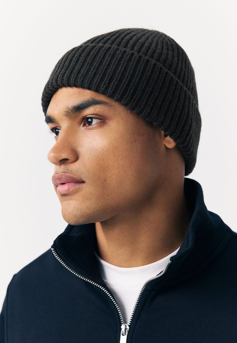 Dark ribbed knit beanie, round shape, worn above the forehead; accompanied by a dark zip-up jacket and white t-shirt underneath.