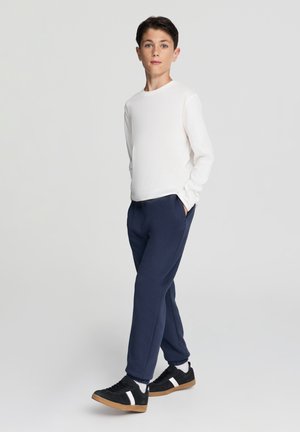 Camicia bianca a maniche lunghe, pantaloni della tuta blu scuro, scarpe da ginnastica nere con dettagli bianchi. Design semplice, texture morbida, vestibilità rilassata.