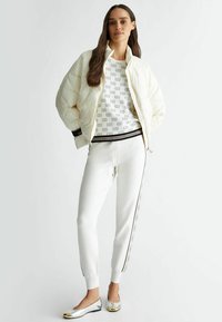 Giacca bianca soffice sopra un maglione bianco a fantasia e pantaloni jogger bianchi con dettagli laterali; ballerine argentate lucide con punte dorate.