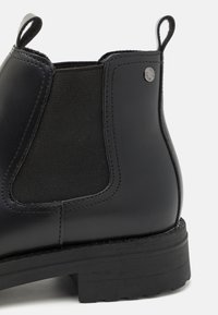 Botte cheville en cuir noir avec panneau latéral élastique, semelle en caoutchouc plate et languette. Présente une couture discrète et un petit logo.