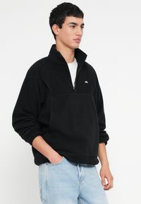 Czarny polarowy pullover z półzamkiem przy kołnierzu i długimi rękawami. Posiada małe logo na piersi oraz luźny krój. Noszony z jasnoniebieskimi dżinsami.