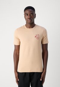 Beige Baumwoll-T-Shirt mit kurzen Ärmeln, das ein rotes Logodesign auf der linken Brust trägt. Schlichte, taillierte Form, glatte Textur und Rundhalsausschnitt.