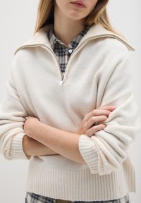 Cremfarbener Reißverschluss-Pullover mit gerippten Bündchen und Saum, aus weichem Stoff gefertigt, über einem karrierten Hemd mit Knöpfen getragen.