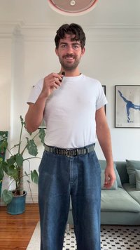 Άνδρας με σκούρα μαλλιά που φοράει ένα λευκό μπλουζάκι και μπλε τζιν κρατάει ένα μικρόφωνο σε ένα σαλόνι με φυτά και καδραρισμένα έργα τέχνης.