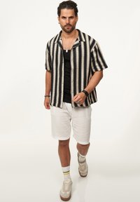 Kurzärmliges Hemd mit Kragen in schwarz-creamfarbenen vertikalen Streifen, kombiniert mit weißen Leinen-Shorts und beigen Sneakers mit grün- und gelbgestreiften Socken.