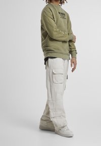 Olivgrön sweatshirt med rund hals och logotyp framtill, tillsammans med löst sittande, beige cargobyxor med flera fickor. Vita sneakers.