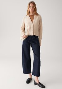 Beige Strickjacke aus Wollmischung mit V-Ausschnitt, fünf Knöpfen und geripptem Saum; kombiniert mit dunkelblauen, weit geschnittenen Jeans und schwarzen Slippern.