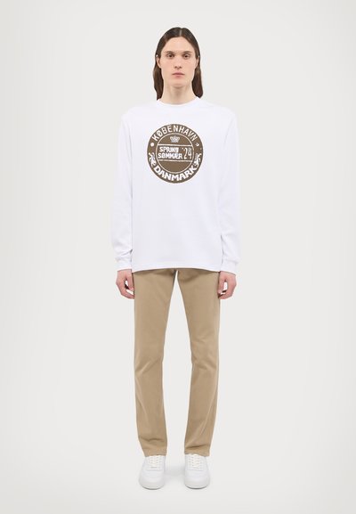 White long-sleeve shirt with round graphic print, beige trousers, and white sneakers. The shirt features a textured fabric and casual fit.