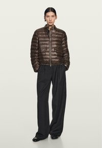 Brown puffer jacket with a high collar, horizontal seams, front zipper, and two side pockets, paired with loose black trousers.