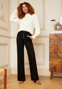 Jersey blanco de punto con patrón texturizado, combinado con pantalones de tiro alto negros de campana que cuentan con cinco botones y bolsillos laterales.
