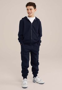 Temno modra bombažna zip-up kapuca z sprednjimi žepe, v kombinaciji z ujemajočimi se trenirkami z bočnimi žepi in elastičnimi manšetami. Bele teniske.