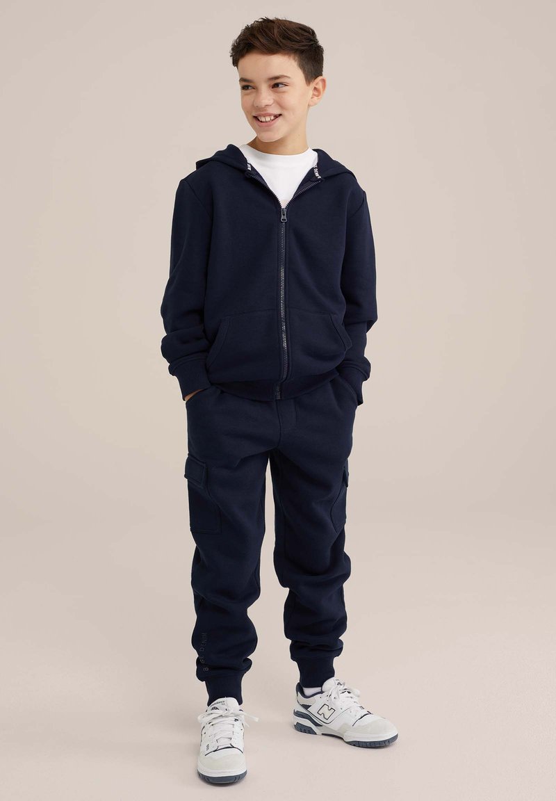 Temno modra bombažna zip-up kapuca z sprednjimi žepe, v kombinaciji z ujemajočimi se trenirkami z bočnimi žepi in elastičnimi manšetami. Bele teniske.