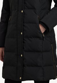 Melnā pūkā, kurai ir quilted dizains, zelta krāsas pogas, sānu kabatas ar rāvējslēdzēju un ribotas aproces papildu siltumam.