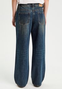 Wijde denim jeans in mediumblauw, met vervaagde plekken, twee achterzakken en een lichte textuur. Leren taillebandlabel zichtbaar aan de achterkant.