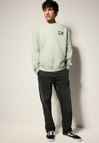 Helles grünes Sweatshirt mit gesticktem Design auf der Brust, kombiniert mit dunklen grauen Jeans und schwarzen Sneakers mit weißen Schnürsenkeln.