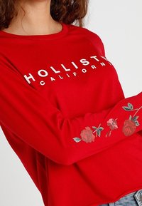 Röd långärmad skjorta med "HOLLISTER CALIFORNIA" i vita bokstäver. Har blommönster på ärmarna med rosor och gröna blad.