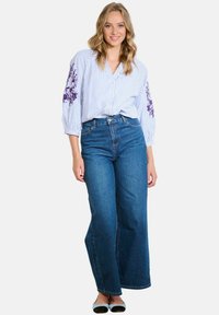 Femme debout portant un chemisier à rayures blanches et bleues avec une broderie florale violette sur les manches, un jean bleu à jambes larges et des ballerines bicolores.