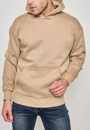 Homme portant un sweat à capuche beige et un jean bleu, les mains près de la poche avant, sur un fond clair.