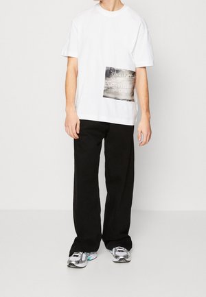 Valkoinen puuvillainen T-paita, jossa on vasemmalla puolella suorakaiteen muotoinen graafinen painatus, yhdistetty mustiin leveälahkeisiin housuihin ja harmaisiin tennareihin.