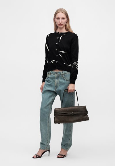 Femme portant un cardigan noir à motifs, un jean bleu clair et des sandales à talons noires, tenant un grand sac à main marron foncé par sa anse.