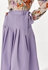 Högmidjad lavendelfärgad kjol med veck och knytbälte, i kombination med en blommig blus med långa ärmar i orange och lila blommor.